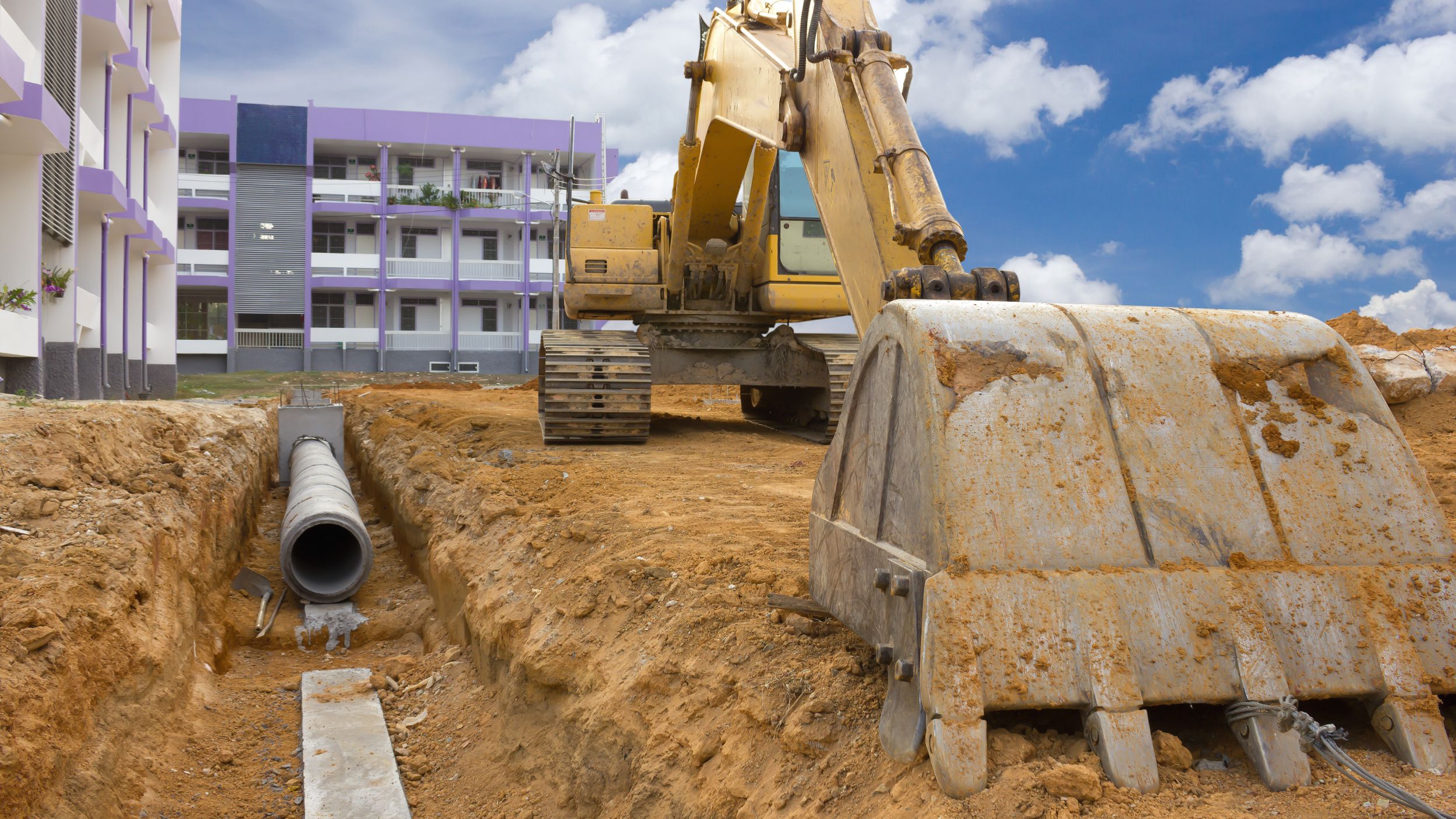 Kettenbagger auf Baustelle beim Aushub eines Grabens mit verlegtem Abwasserrohr vor modernem Wohngebäude